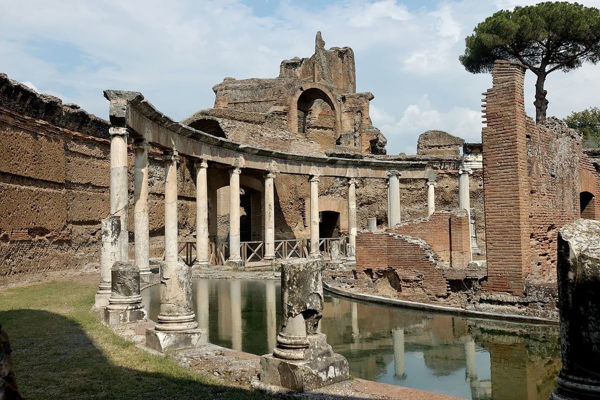 Ruins of the ancient Roman villa at Hadrian's Villa in Tivoli, Italy, featuring a circular colonnade surrounding a tranquil reflecting pool, with remnants of brick walls and trees in the background under a blue sky.