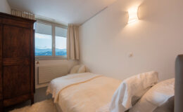 Cozy bedroom featuring a single bed and a double bed with white bedding, an antique wooden wardrobe, and large windows offering scenic mountain views. Soft lighting creates a warm and inviting atmosphere.
