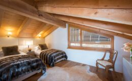 Cozy attic bedroom featuring two twin beds with plush bedding, wooden beams, and a large window offering natural light, complemented by a stylish chair and decorative accents. Ideal for a relaxing retreat.