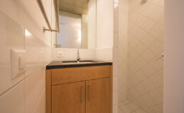 Modern bathroom interior featuring a wooden vanity with a black countertop, sink, and a glass-enclosed shower with tiled walls. Natural light from a window enhances the contemporary design.