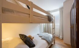 Cozy children's bedroom featuring a white bunk bed with a lower twin bed, plush pillows, and a teddy bear, illuminated by natural light from a window.