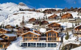 Charming alpine village with wooden chalets surrounded by snow-covered mountains under a clear blue sky, ideal for winter vacations and skiing.