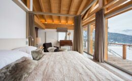 Modern bedroom with wooden beams, large windows offering mountain views, and a cozy bed adorned with soft bedding and decorative pillows. The room features a minimalist design with warm tones, a comfortable seating area, and access to a balcony.
