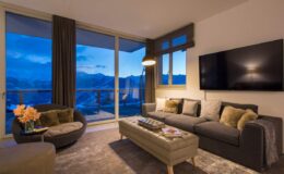 Modern living room with large windows showcasing mountain views, featuring a comfortable gray sofa, decorative pillows, a stylish coffee table, and a flat-screen TV, illuminated by warm lighting in the evening.