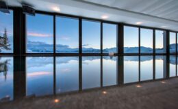 Modern indoor swimming pool with panoramic mountain views, featuring large glass windows that reflect the serene landscape at dusk.