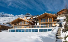 Modern wooden chalet in a snowy mountain landscape, featuring large windows and a spacious terrace, surrounded by pristine snow and scenic views.