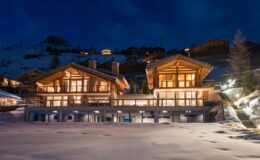 Luxury chalet in a snowy landscape at night, featuring large windows and wooden architecture, surrounded by mountains. Perfect for winter retreats and upscale vacations.