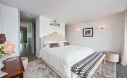 Modern hotel bedroom featuring a king-size bed with white bedding, decorative throw pillows, and a stylish bench at the foot. The room is elegantly designed with soft lighting, neutral tones, and a plush area rug, creating a cozy and inviting atmosphere.