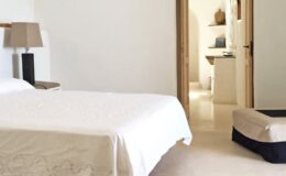 Modern bedroom with a white bedspread, a bedside lamp, and an open doorway leading to a minimalist bathroom. The decor features neutral tones and simple furnishings, creating a serene atmosphere.