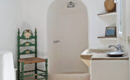 Charming minimalist bathroom featuring a whitewashed wall, a green wooden chair, a stone sink, and a shower alcove, enhancing the serene and rustic ambiance.