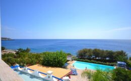 Scenic view of a coastal landscape featuring a clear blue sea, lush greenery, and a swimming pool under a bright sky. Ideal for vacation rentals or travel destinations.