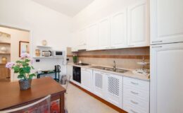 Modern kitchen interior featuring white cabinetry, a wooden dining table with a flower centerpiece, and essential appliances including a microwave and kettle. The design emphasizes a clean and functional layout with ample counter space.