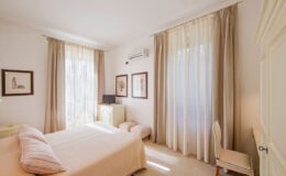 Bright and airy bedroom featuring a light-colored bedspread, large windows with sheer curtains, and tasteful artwork on the walls. The room includes a small dresser and a comfortable chair, creating a cozy and inviting atmosphere.