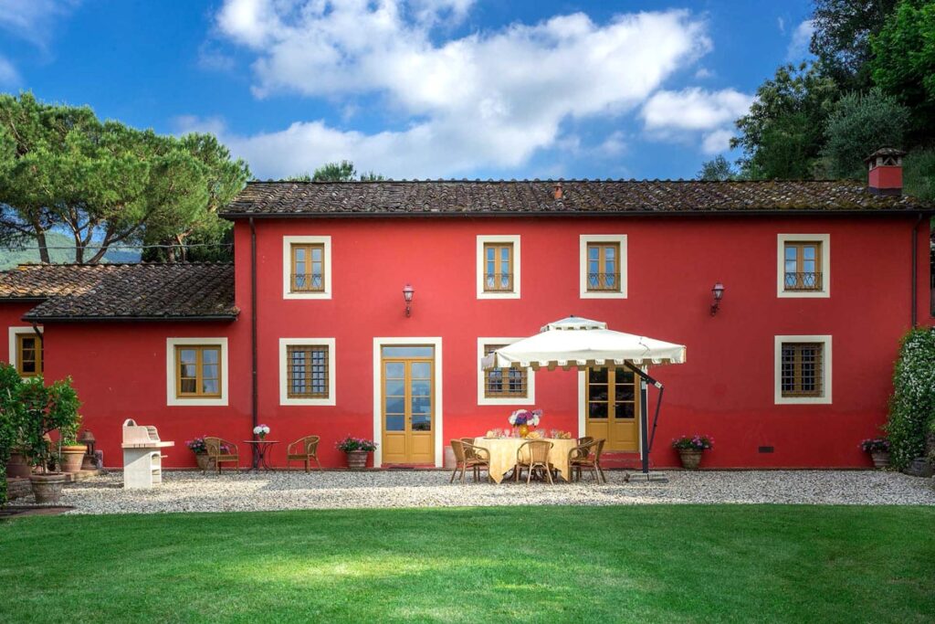 Charming red villa with a patio and outdoor dining area surrounded by lush greenery and blue skies, ideal for a peaceful getaway. | Only on SopranoVillas