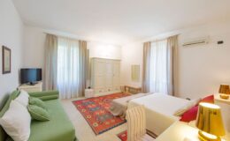 Modern hotel room featuring a cozy green sofa, a double bed with white linens, a colorful area rug, and large windows with sheer curtains. The space includes a flat-screen TV, a wardrobe, and stylish lighting, creating a bright and inviting atmosphere.