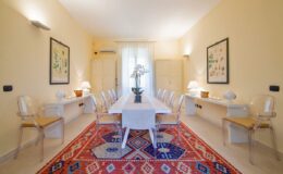 Bright and airy dining room featuring a long white table surrounded by transparent chairs, complemented by a colorful patterned rug and decorative wall art. Natural light filters through large windows, enhancing the inviting atmosphere.