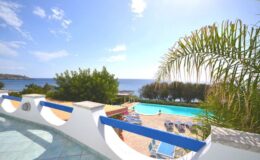 Scenic view from a balcony overlooking a tranquil pool area and the ocean, surrounded by lush greenery and clear blue skies. Ideal for vacation rentals or travel destinations.