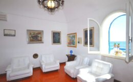 Bright living room with white furniture, artistic wall decor, and a large window offering a scenic view of the sea. The space features a chandelier and warm, terracotta flooring, creating a cozy and inviting atmosphere.