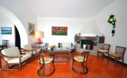 Cozy living room featuring a fireplace, comfortable seating with wooden chairs, a coffee table, and colorful artwork on the walls. The bright orange tiled floor adds warmth to the space, complemented by soft lighting from a stylish lamp and wall sconces.