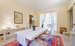 Bright and inviting dining room featuring a large white wooden table, clear acrylic chairs, and a colorful patterned rug. Natural light streams through sheer curtains, highlighting decorative elements like a potted orchid and wall art.