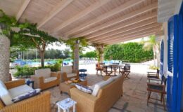 Cozy outdoor living space featuring wicker furniture, a wooden ceiling, and lush greenery, ideal for relaxation and entertaining by the poolside.