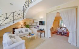 Bright and airy living room featuring a white sofa, decorative shelves, and a flat-screen TV, with a staircase leading to an upper level and an open doorway to a dining area.