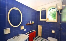 Bright blue tiled bathroom featuring a round mirror, a window with a view, a small shelf with toiletries, a towel rack with red towels, and a decorative radiator.