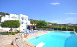 Stunning white villa with blue accents, surrounded by lush greenery, featuring a crystal-clear swimming pool and sun loungers under a bright blue sky. Ideal for luxury vacation rentals.