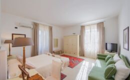 Spacious and elegantly designed hotel room featuring a comfortable bed, green seating area, wooden furniture, and large windows with natural light, ideal for relaxation and leisure.