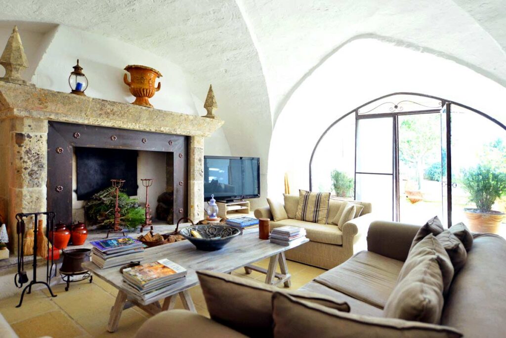 Cozy living room with an arched ceiling, featuring a stone fireplace, a large TV, and comfortable seating. Natural light streams in through a large window, and decorative elements include plants and books on the coffee table. | Only on SopranoVillas