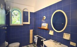 Bright blue tiled bathroom featuring a round mirror, a window with an arched design, a white sink, and a toilet. The space includes a towel rack and decorative elements, creating a serene and stylish atmosphere.