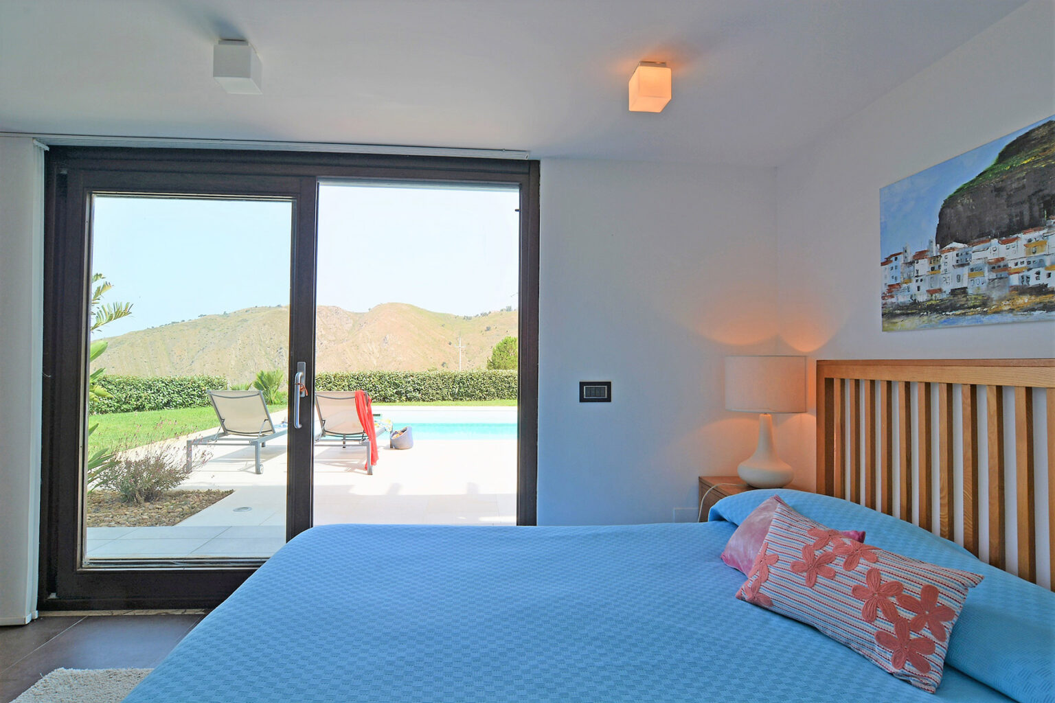 Bright and modern bedroom featuring a blue bedspread, coral accent pillows, and large glass doors leading to an outdoor pool area with lounge chairs and scenic mountain views. | Only on SopranoVillas