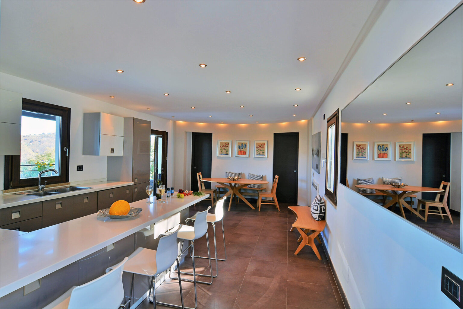 Modern kitchen with sleek cabinetry, a spacious island, and contemporary seating. Features large windows for natural light, a dining area with wooden furniture, and stylish lighting fixtures. Ideal for entertaining and cooking. | Only on SopranoVillas