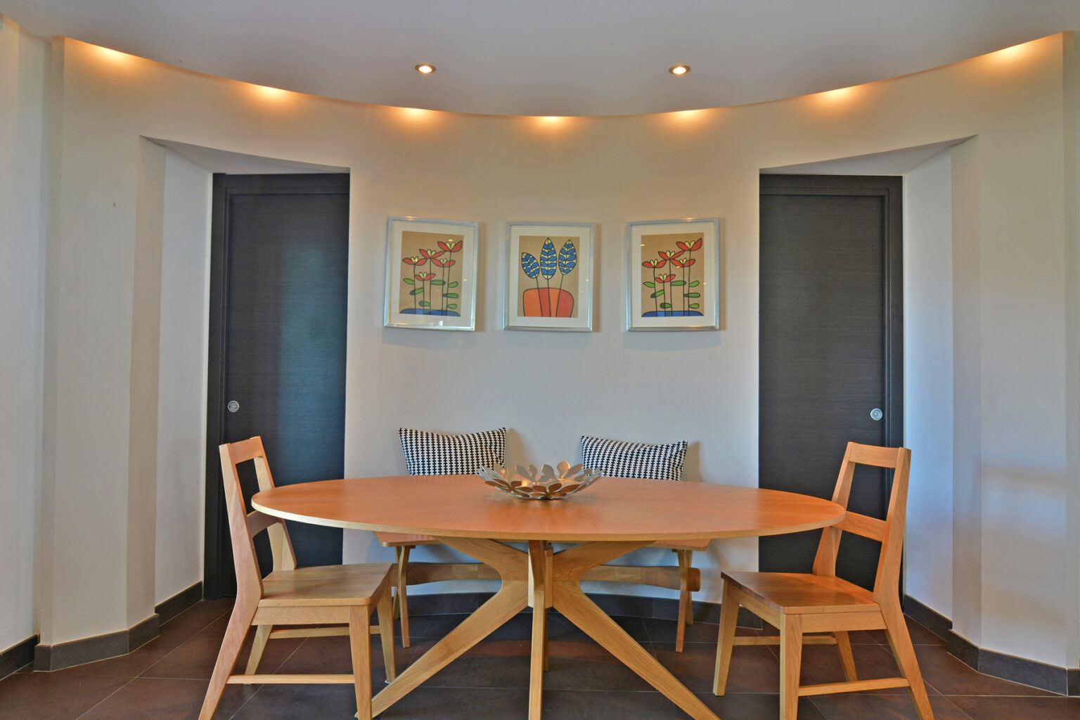 Modern dining area featuring a round wooden table and two wooden chairs, complemented by decorative wall art and soft lighting. The design emphasizes a cozy and inviting atmosphere. | Only on SopranoVillas