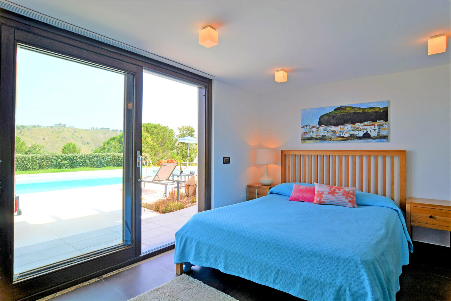 Bright and airy bedroom featuring a wooden bed with a blue blanket, large windows overlooking a pool and garden, and a colorful painting on the wall. Ideal for relaxation and comfort in a modern home. | Only on SopranoVillas