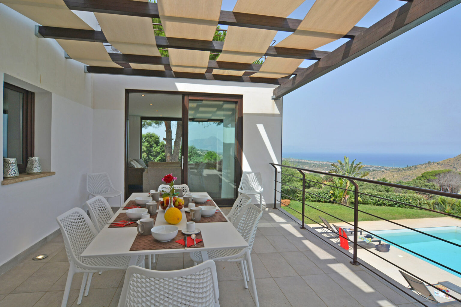 Outdoor dining area with a modern table set for a meal, surrounded by white chairs, overlooking a scenic view of a pool and lush greenery under a pergola. Ideal for enjoying meals in a tranquil setting. | Only on SopranoVillas