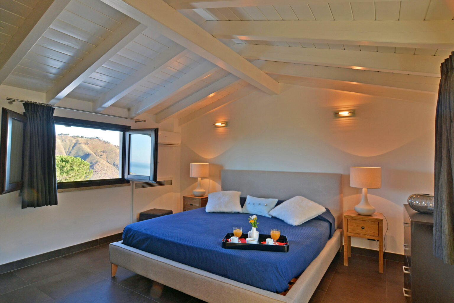 Cozy bedroom with a blue bedspread, two bedside lamps, and large windows offering scenic views. The room features wooden beams and modern decor, creating a serene atmosphere. Perfect for relaxation and comfort. | Only on SopranoVillas