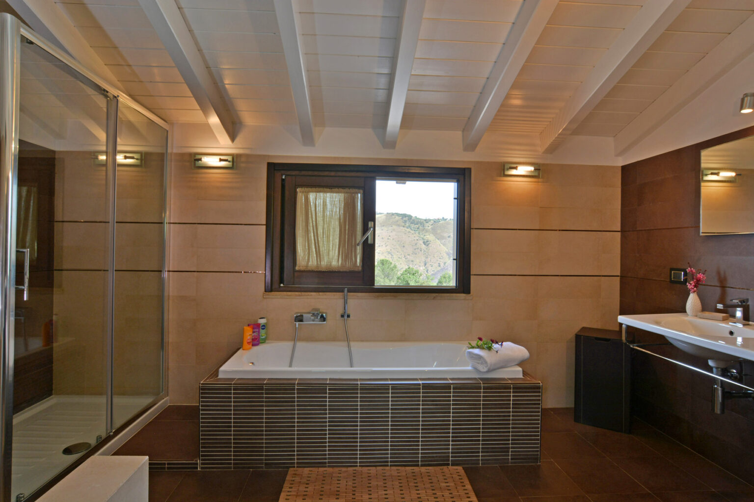 Modern bathroom interior featuring a spacious bathtub, glass shower enclosure, and stylish wooden accents. Natural light flows through a window, enhancing the contemporary design. | Only on SopranoVillas