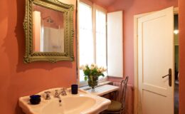Charming bathroom interior featuring a vintage green-framed mirror, a white sink with elegant fixtures, a small table with a vase of fresh flowers, and warm peach-colored walls. Natural light filters through the windows, enhancing the cozy atmosphere.