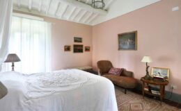 Cozy bedroom featuring a double bed with white linens, a comfortable armchair, and elegant decor including framed artwork on pastel pink walls. Natural light floods the space through large windows, enhancing the inviting atmosphere.