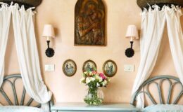 Cozy bedroom decor featuring a light pink wall, two twin beds with blue headboards, white sheer curtains, wall-mounted sconces, and a floral arrangement in a vase on a bedside table, complemented by decorative wall art and framed pictures.