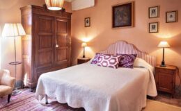 Cozy bedroom featuring a double bed with a white coverlet, decorative pillows, antique wooden wardrobe, bedside lamps, and framed artwork on peach-colored walls, creating a warm and inviting atmosphere.