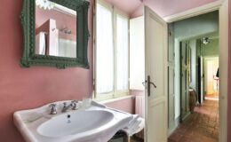 Elegant bathroom interior featuring a vintage sink and a green-framed mirror, with natural light streaming through large windows and a view of a hallway in soft pastel colors.