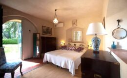 Cozy bedroom featuring a double bed with decorative pillows, elegant lighting, and a vintage wooden dresser. A large window offers a view of a lush garden, enhancing the room's inviting atmosphere. Ideal for relaxation and comfort.