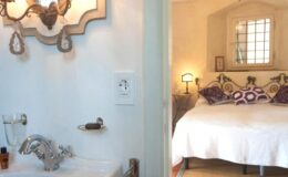 Cozy bedroom with a double bed and decorative pillows, visible through an open doorway, alongside a stylish bathroom featuring a modern sink and elegant wall sconces.