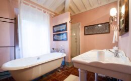 Elegant bathroom featuring a freestanding bathtub, vintage sink, and natural light from large windows, adorned with pastel pink walls and rustic wooden beams.