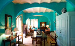Bright and airy living room featuring turquoise walls, arched ceilings, wooden dining table, and cozy seating. The space is illuminated by natural light, complemented by decorative lamps and artwork, creating a welcoming atmosphere.