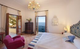 Spacious bedroom with a sea view, featuring a comfortable bed, elegant chandelier, and a cozy armchair. The room has large windows that open to a balcony, allowing natural light to fill the space. Decorated with stylish furnishings and tasteful accents.