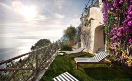 Scenic terrace with striped lounge chairs overlooking the ocean, surrounded by lush greenery and vibrant purple flowers, ideal for relaxation and enjoying stunning views.