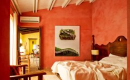 Cozy bedroom with red plaster walls, featuring a wooden bed with white bedding, a large artwork on the wall, and a view into a brightly colored adjoining room. Natural light floods the space, highlighting the rustic decor and inviting atmosphere.